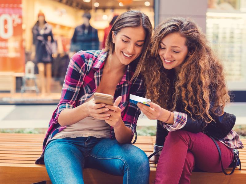 zwei Mädchen sitzen vor einem Geschäft und schauen auf ihre Smartphones